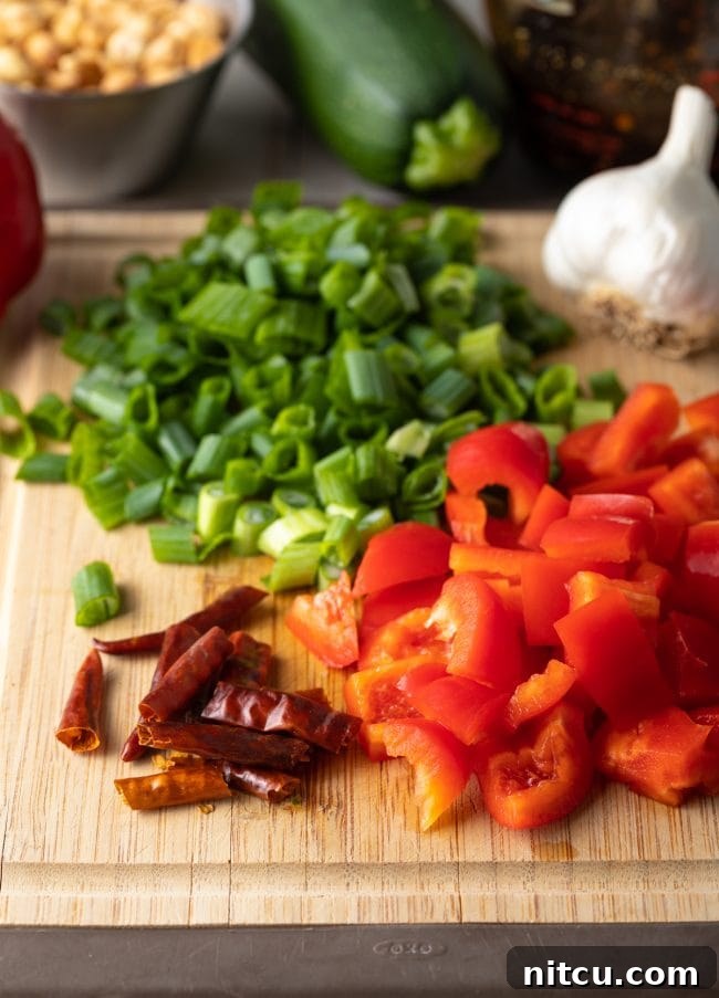 Diced bell peppers, scallions, red chiles and a bulb of garlic on a cutting board.