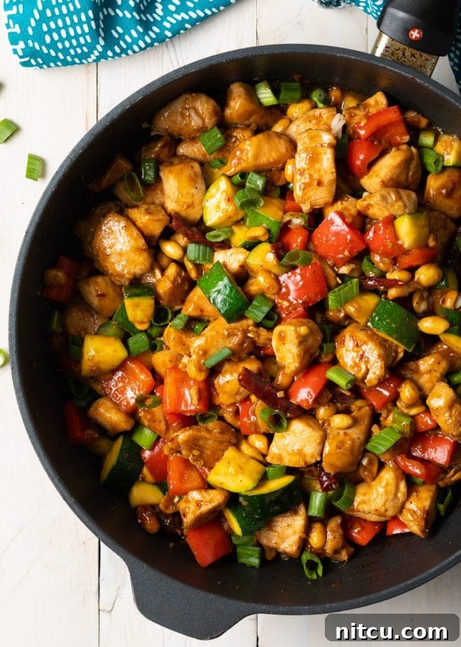 Top down view of a cast iron skillet with chopped cooking chicken, peanuts, chopped scallions, zucchini and bell pepper.