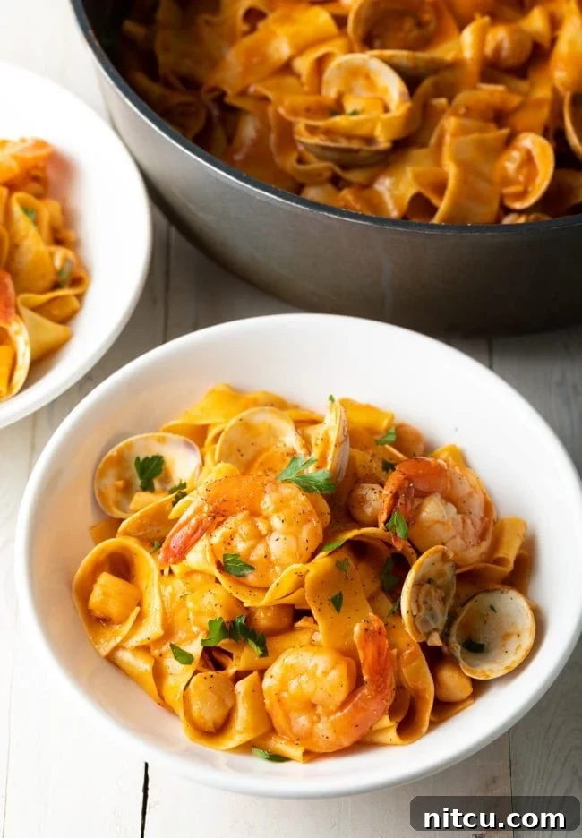 A beautifully plated serving of pappardelle pasta with a rich seafood tomato cream sauce, garnished with fresh parsley in a white bowl.