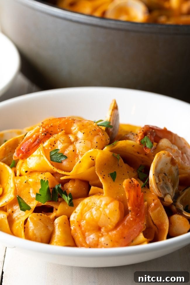 Close-up of a serving of pappardelle pasta coated in a creamy seafood sauce, with visible shrimp and scallops, in a white bowl.
