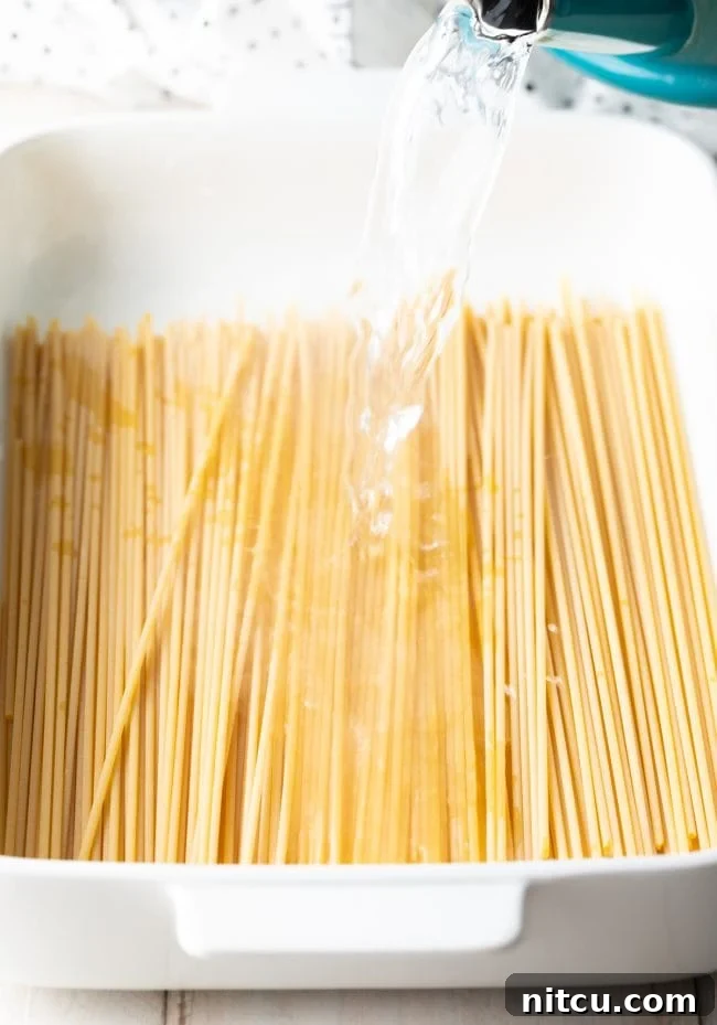 Pouring hot water over bucatini pasta in a white baking dish.
