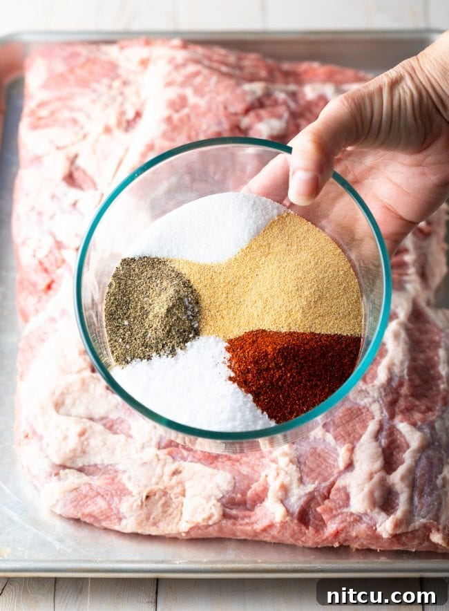 Dry brisket brine spice rub being applied by hand to a raw beef brisket on a baking sheet.