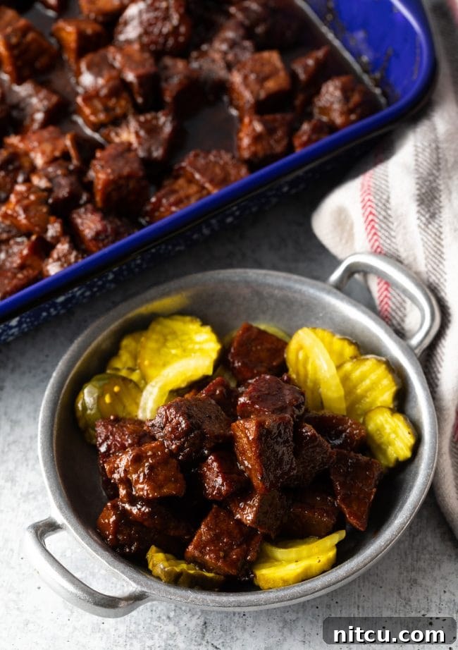 Overhead shot of a metal bowl brimming with smoky, saucy brisket burnt ends, alongside bread and butter pickles.