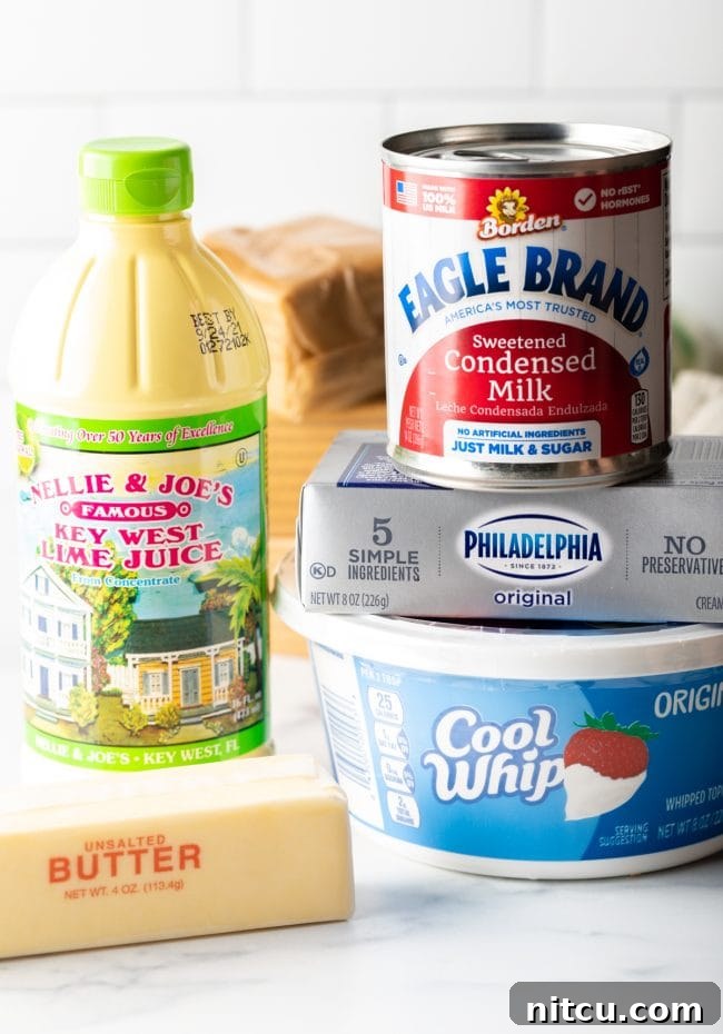 A collection of key ingredients laid out: softened cream cheese, a tub of Cool Whip, bottled key lime juice, sweetened condensed milk, sticks of butter, and a stack of graham crackers, ready for pie making.
