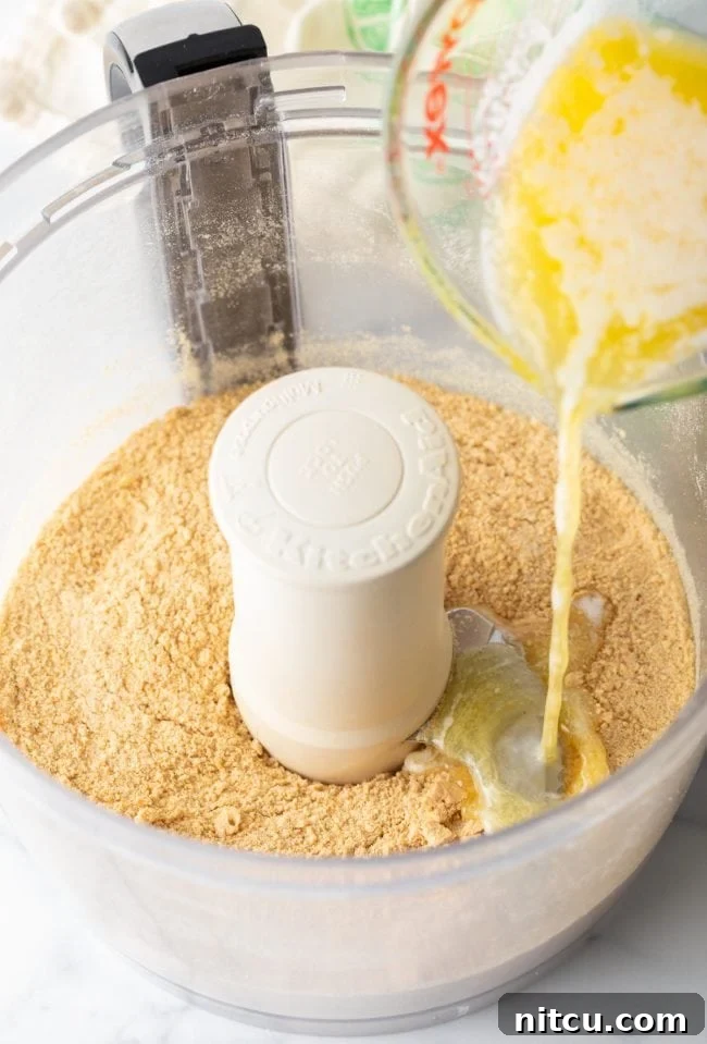 Graham cracker crumbs inside a food processor bowl, with a stream of melted butter being poured over them, ready for blending to form the pie crust.