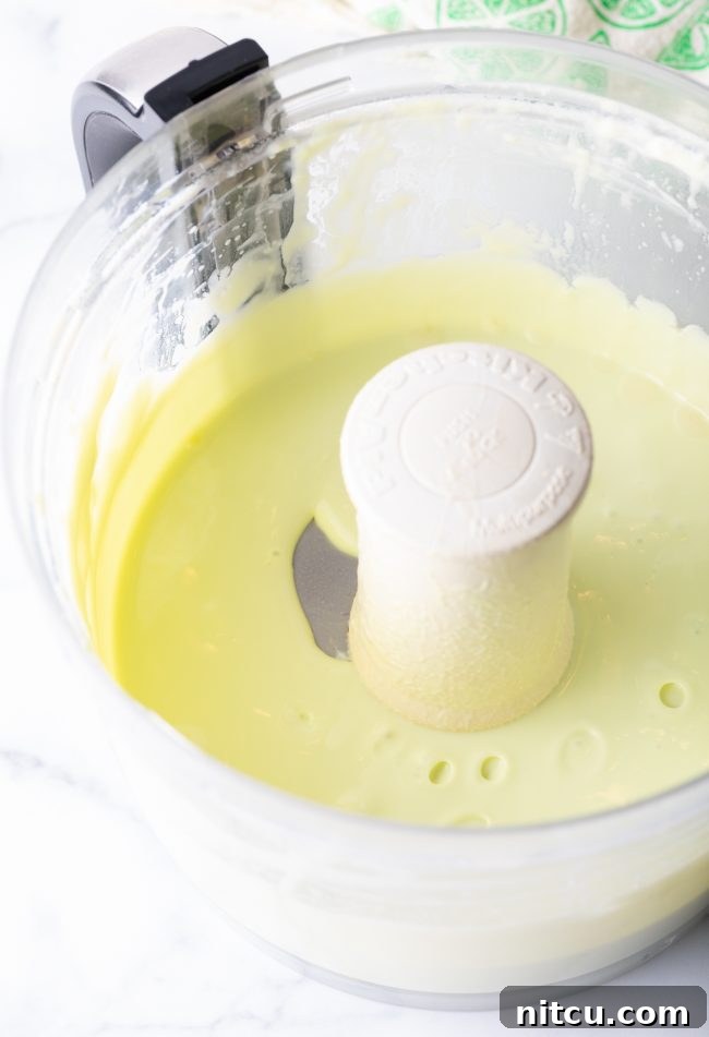 A bright green key lime pie filling mixture, smooth and creamy, swirling inside a food processor bowl, ready for the next step.