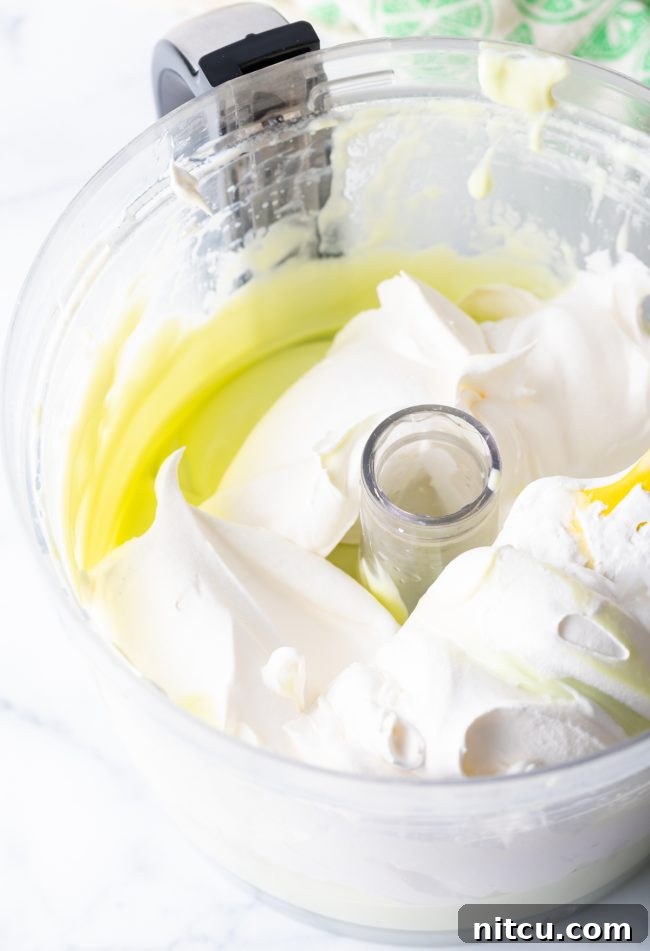 A large scoop of fluffy Cool Whip being added to the bright key lime filling mixture inside a food processor bowl, preparing for the gentle folding process.
