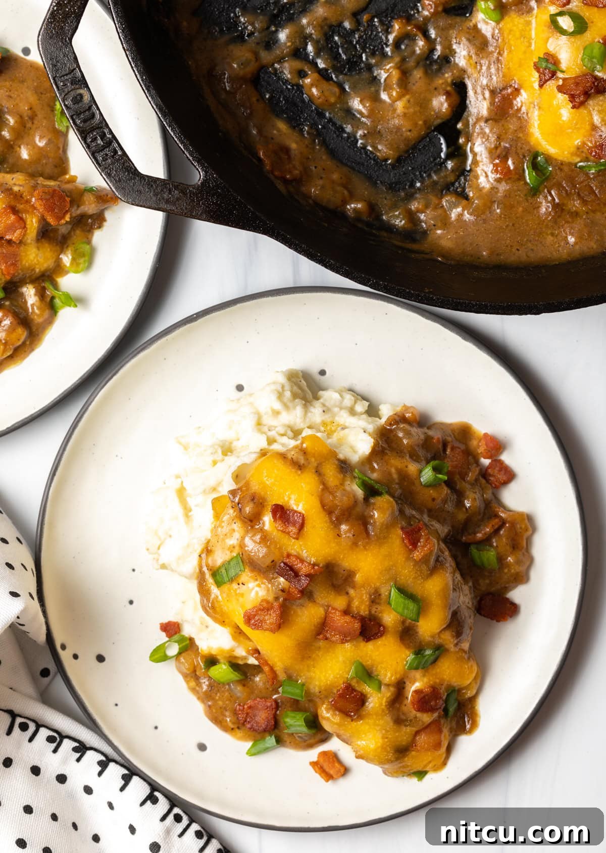 Overhead view of a white plate with fluffy mashed potatoes and a generous serving of smothered chicken with gravy and cheese, with the skillet in the background.