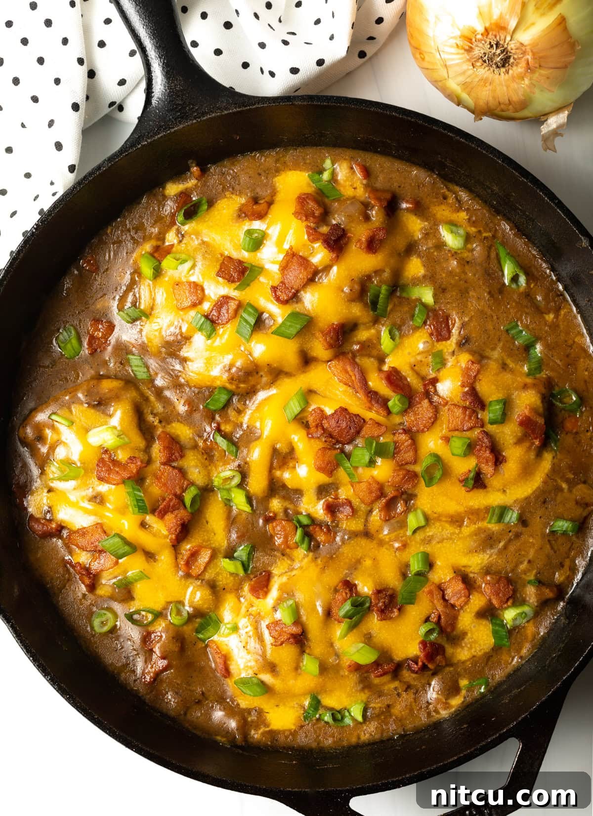 Overhead view of a cast iron skillet filled with golden-brown chicken, rich gravy, melted cheese, crispy bacon, and fresh green onions.