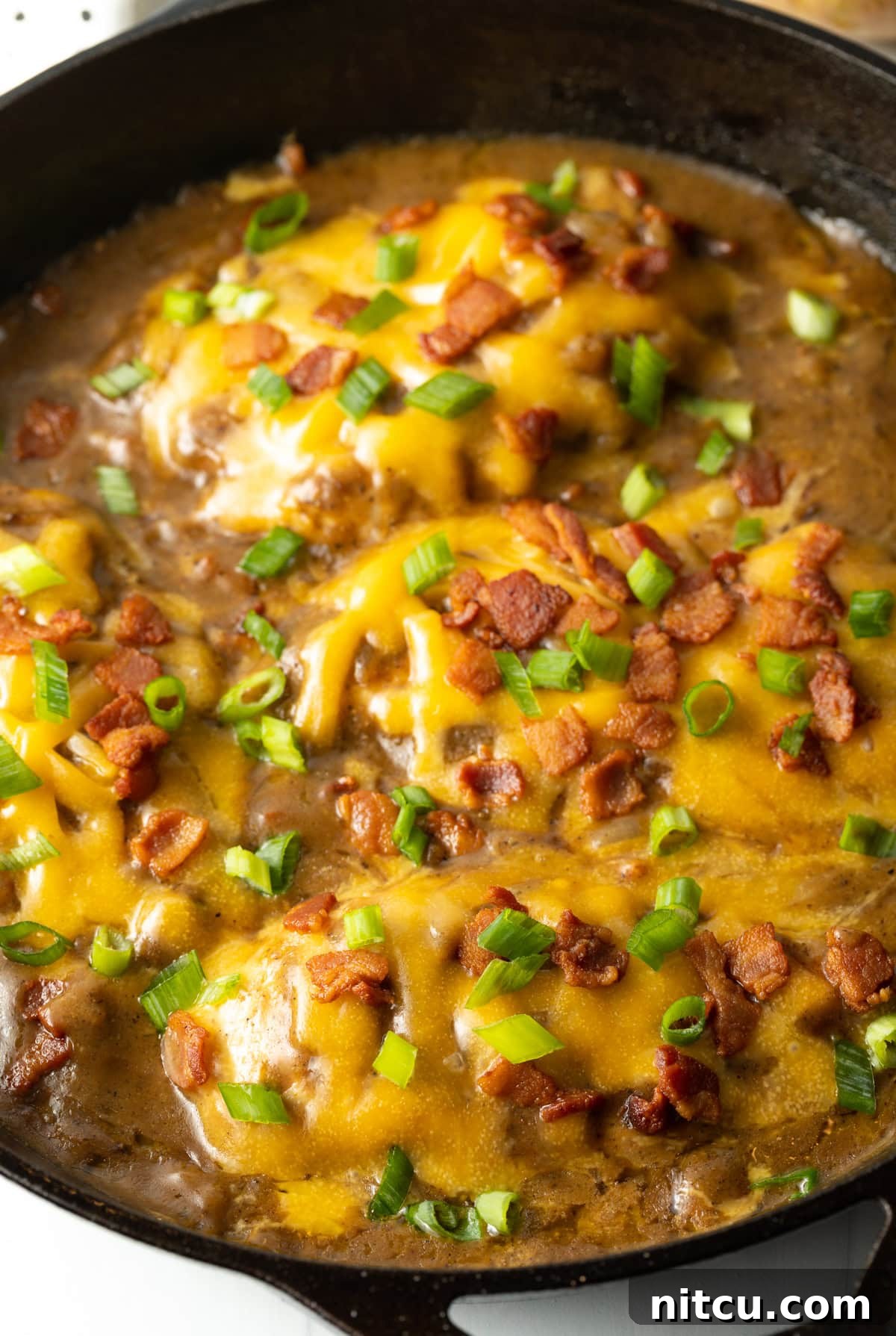 Close up side view of smothered chicken and gravy recipe made with cheese, bacon and scallions in a cast iron skillet, ready to be served.