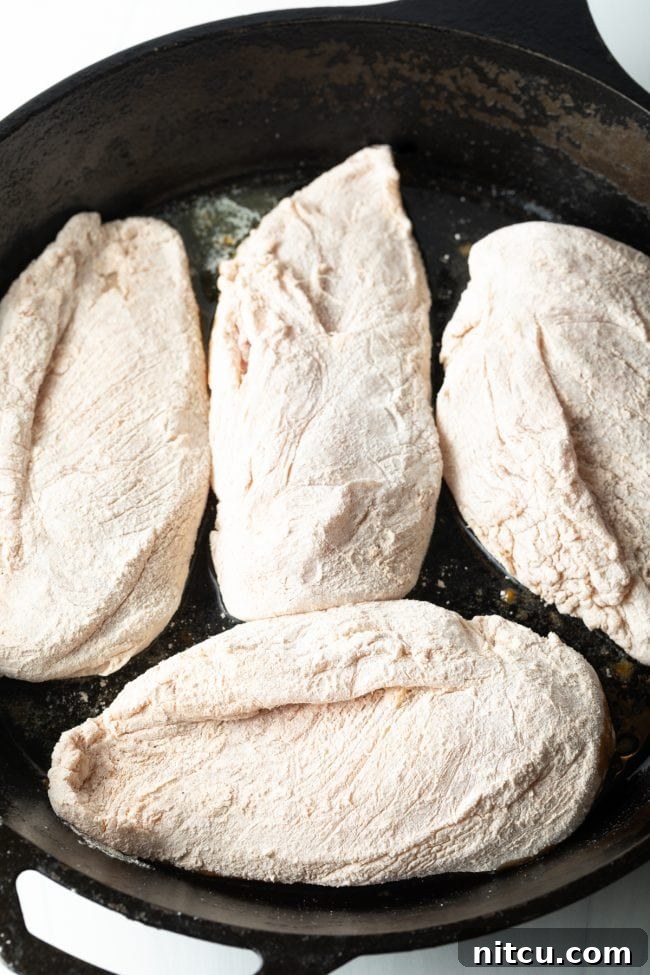 Overhead view of a cast iron skillet holding four flour-coated chicken breasts, nestled in melted butter, ready for the oven.