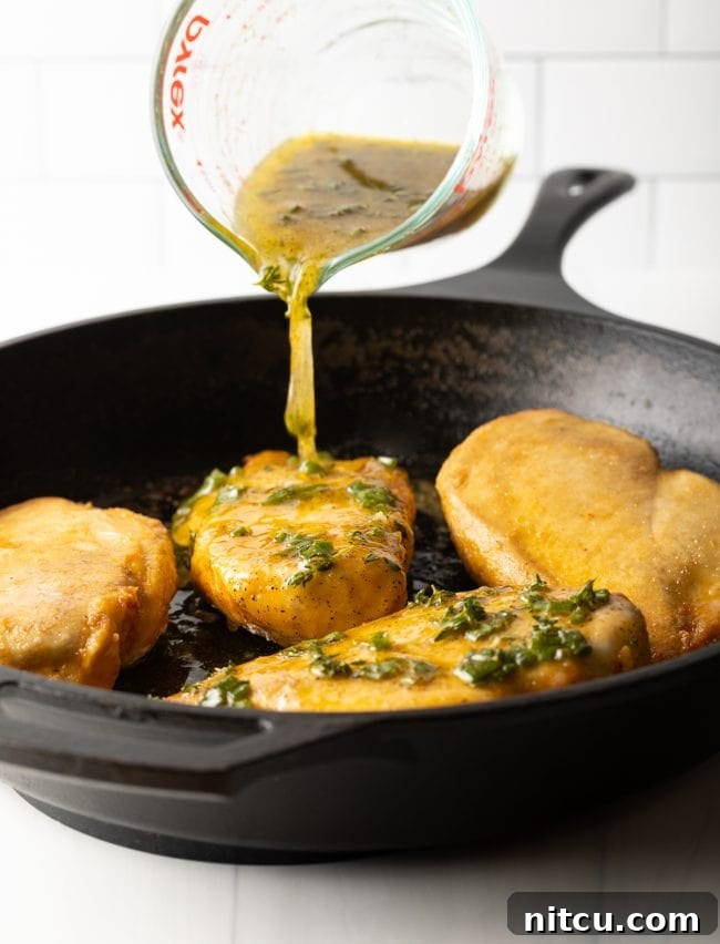 A cast iron skillet filled with golden chicken breasts, ready for the final bake, as vibrant lemon garlic sauce is poured over them from a glass measuring cup.