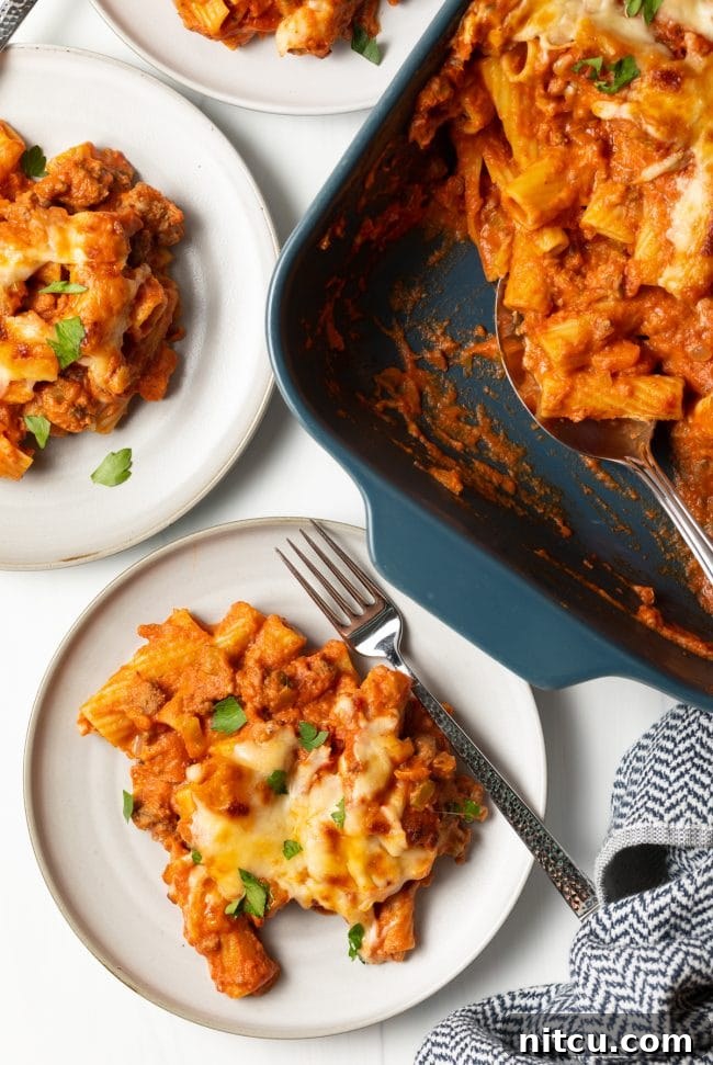 Overhead view of a finished casserole dish of baked rigatoni bolognese, with a serving removed onto a white plate with a fork, ready to be enjoyed.