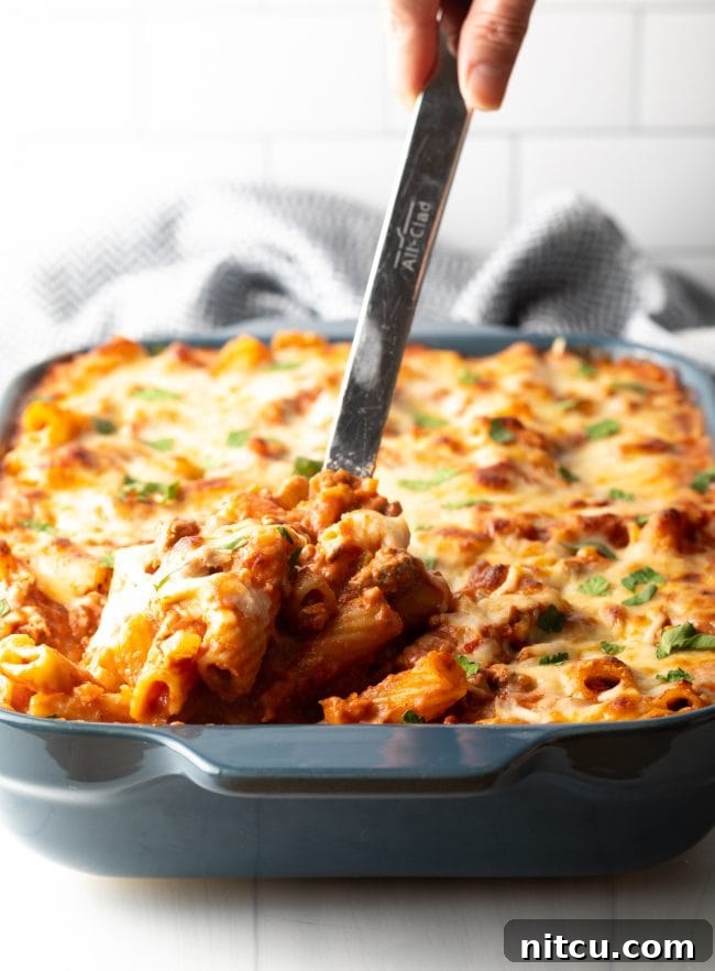 A large metal spoon scoops a perfect serving of cheesy baked pasta with rich bolognese sauce from a casserole dish.