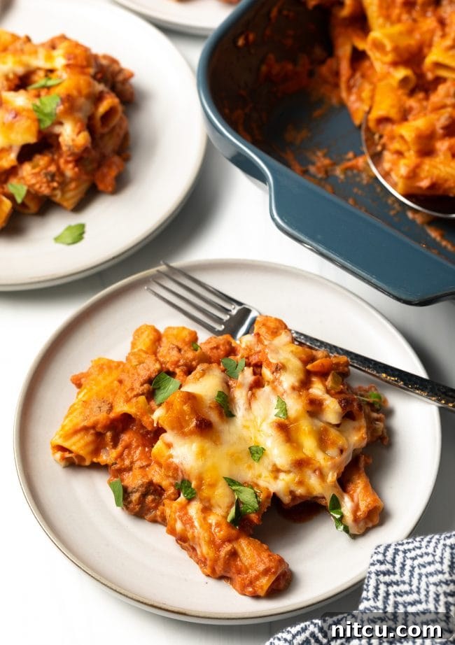 Top-down view of a finished plate of cheesy baked rigatoni bolognese with a fork, with another serving and the casserole dish blurred in the background, inviting indulgence.