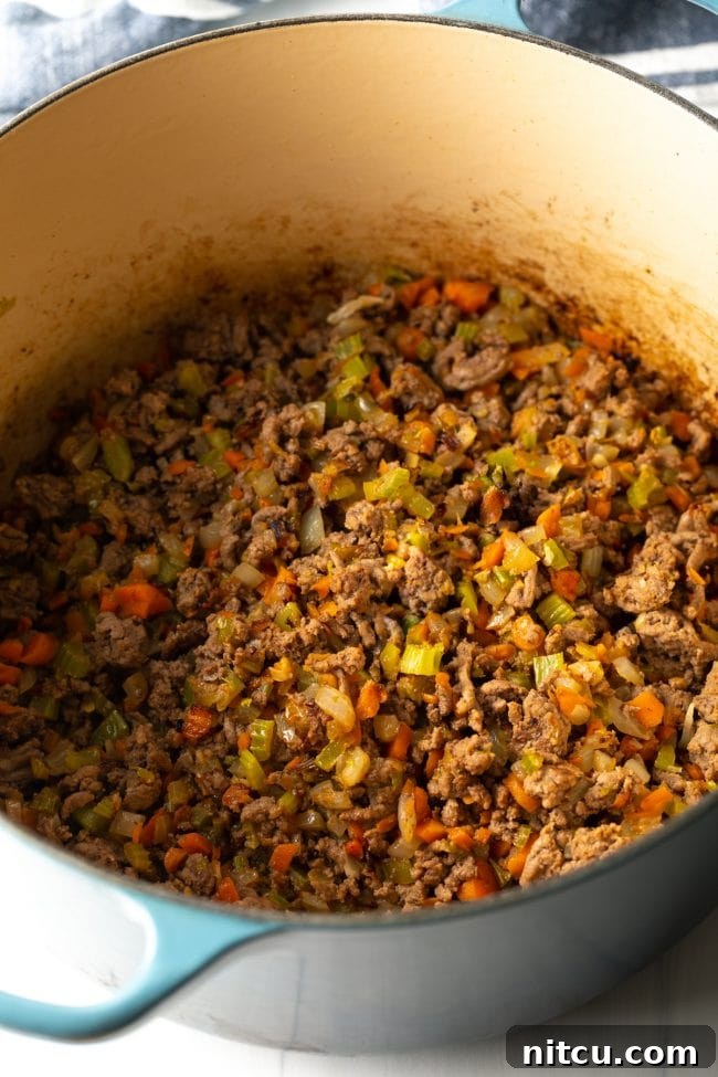 Large Dutch oven on a stovetop, filled with perfectly browned ground beef and sautéed aromatic vegetables, forming the rich base of the bolognese.