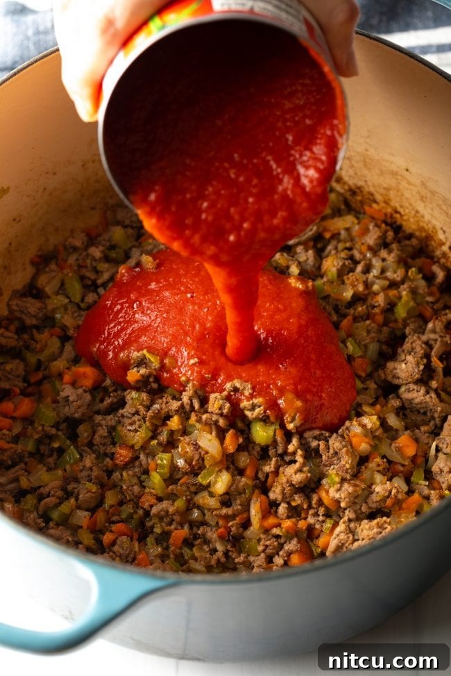 Canned tomato sauce being poured into the large pot containing cooked ground beef and sautéed vegetables, beginning the rich bolognese sauce.