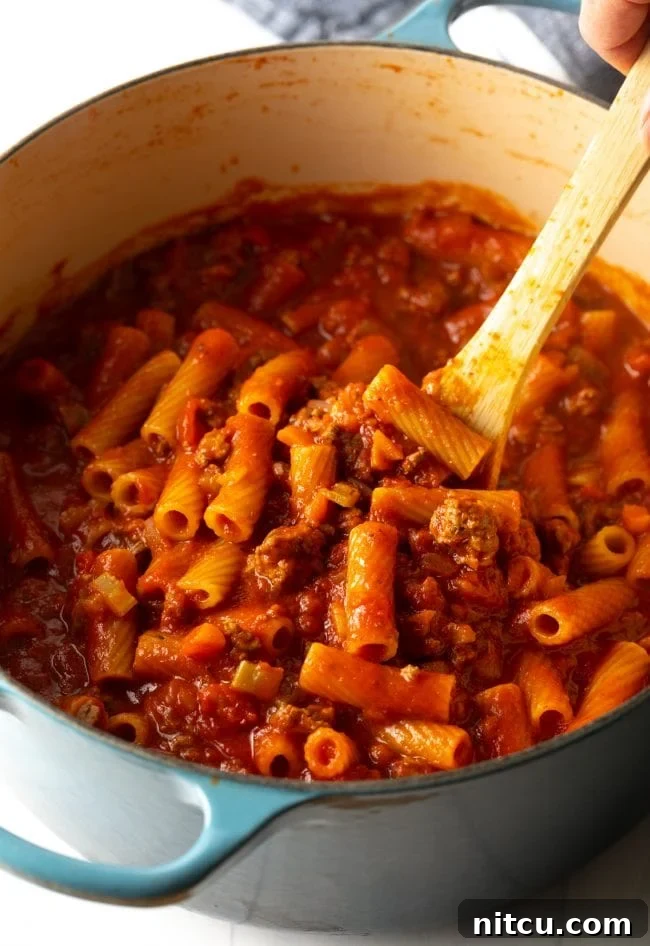 A wooden spoon stirring rigatoni pasta noodles into the simmering tomato, beef, and vegetable mixture, ensuring even cooking and flavor absorption.
