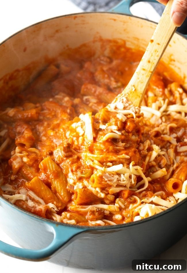 A wooden spoon blending shredded mozzarella cheese and heavy cream into the pasta, tomato, beef, and vegetable mixture, creating a rich and creamy sauce.