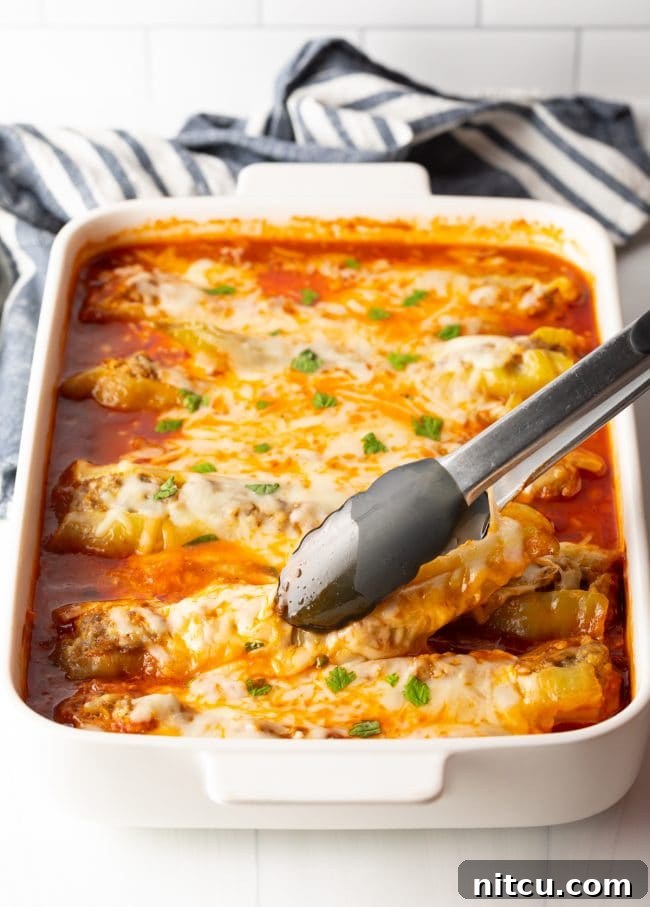 Metal tongs carefully lifting a perfectly stuffed banana pepper, brimming with Italian sausage and cheese, from a baking dish filled with marinara sauce