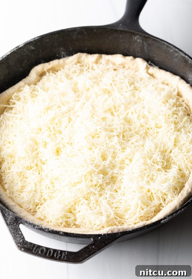 Shredded Italian cheese blend being meticulously spread across the raw pizza dough within a cast iron skillet, forming the essential first layer of the deep dish pizza.