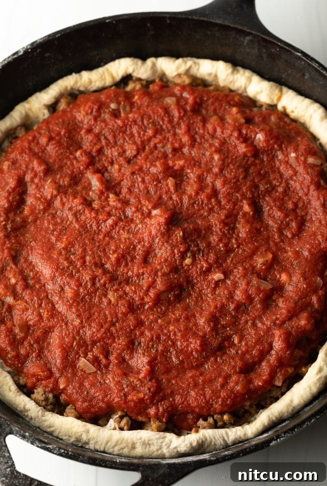 Thick, chunky tomato sauce spread generously over the browned sausage and cheese layers, completing the traditional Chicago deep dish pizza assembly.