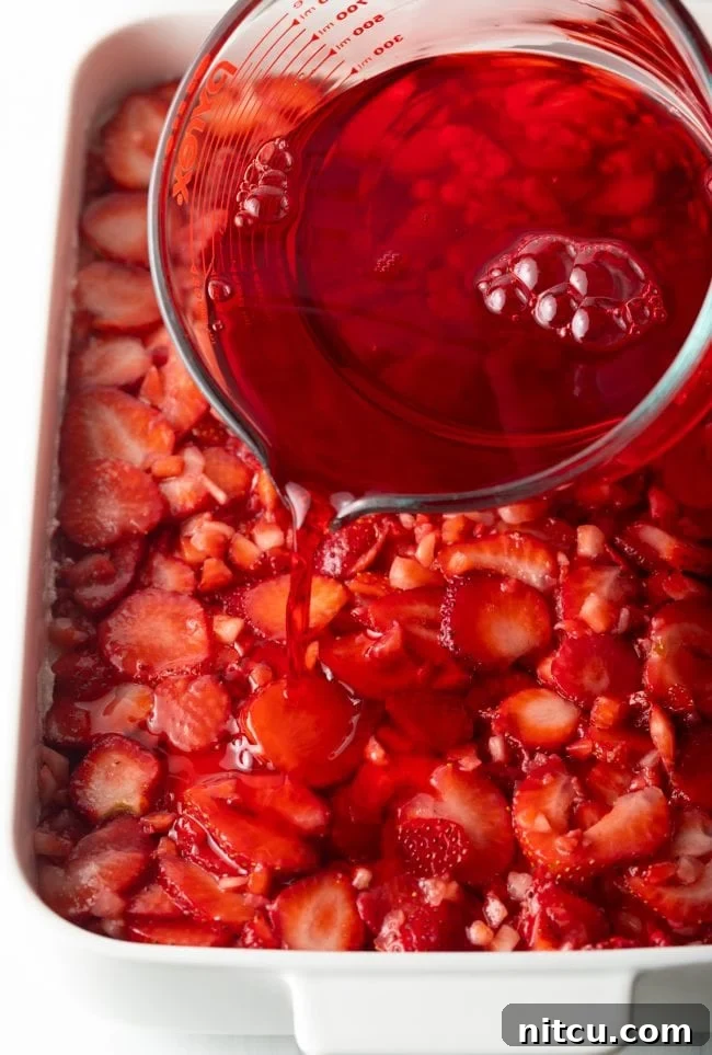 Pouring the cooled red jello mixture over the layers of diced strawberries and cream cheese in the strawberry pretzel dessert recipe.
