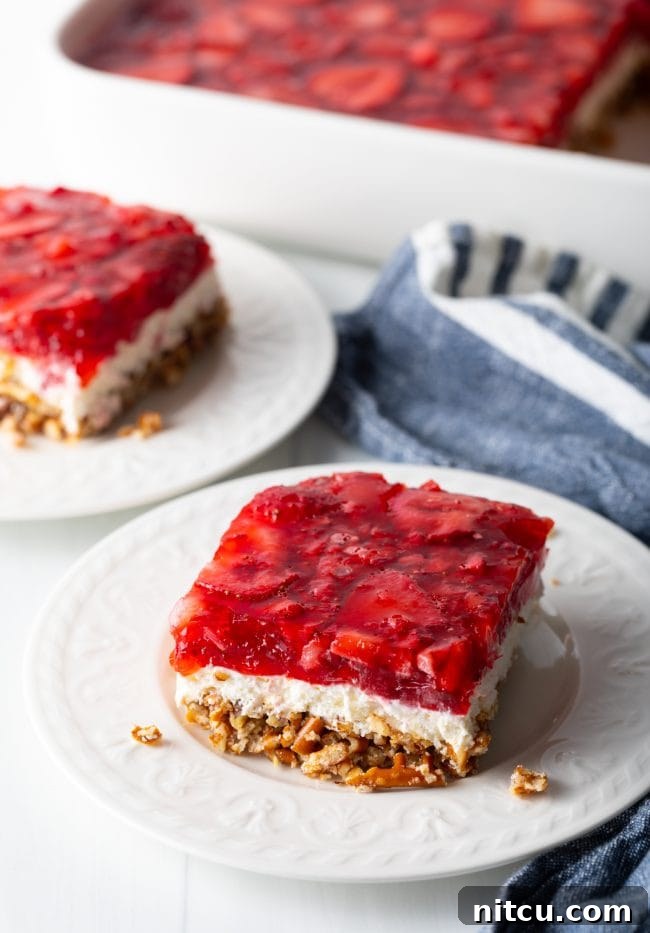 Side view of a perfectly square piece of strawberry pretzel salad recipe on a white plate, showing all three layers.