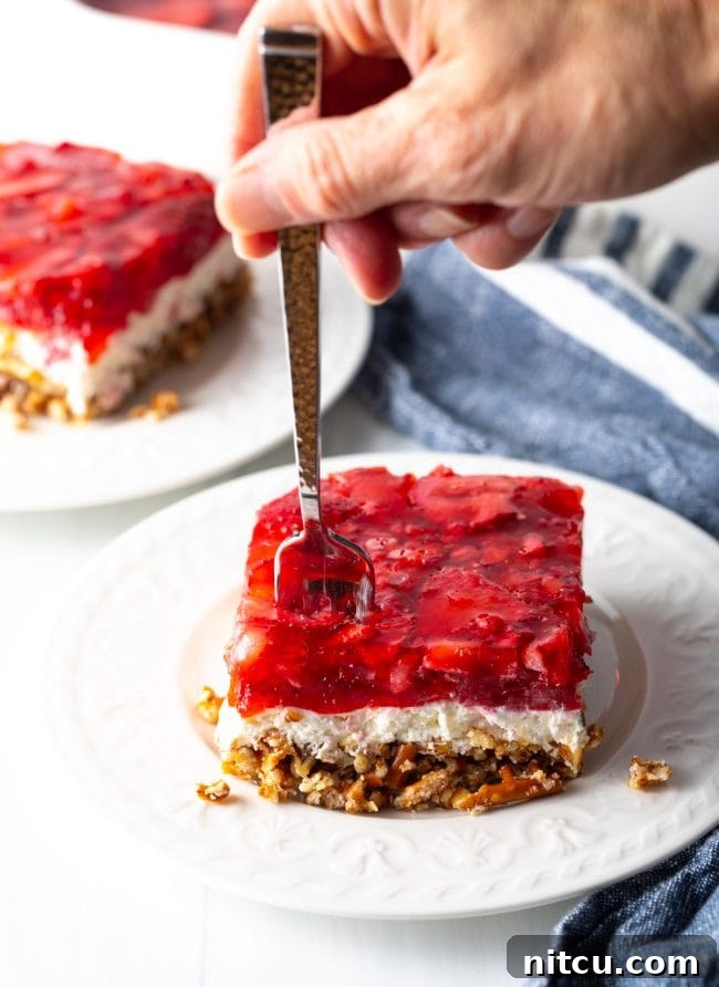 A hand with a fork taking a bite of the layered strawberry jello, cream cheese, and pretzel pecan crust dessert.