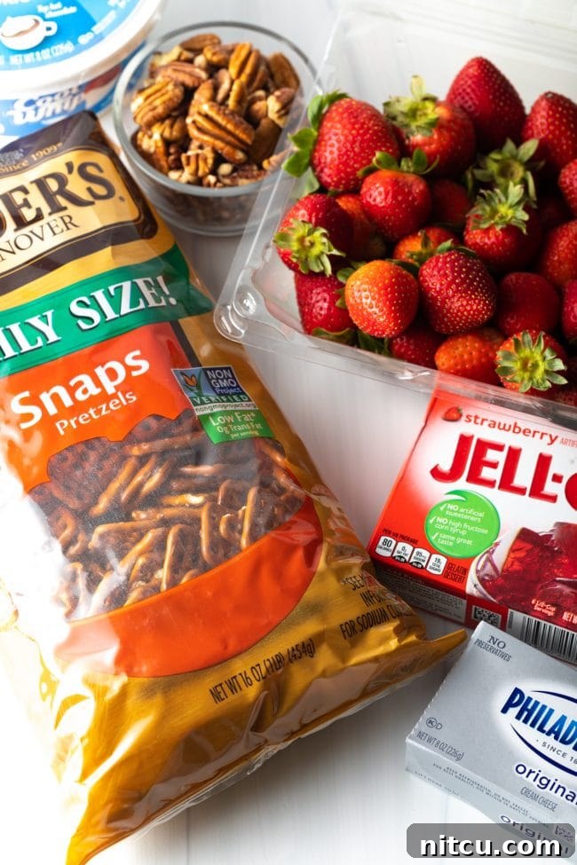 A top-down view of ingredients laid out: a basket of fresh strawberries, a box of strawberry jello, cream cheese, a bag of pretzels, a bowl of pecans, and a tub of cool whip.