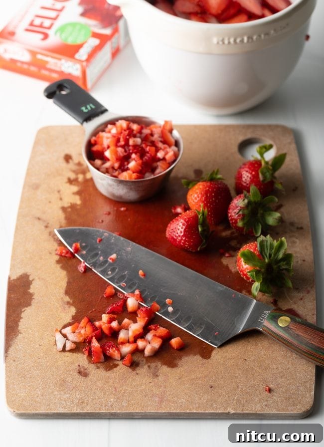 A cutting board with a large knife and both whole and diced strawberries, prepared for the dessert.