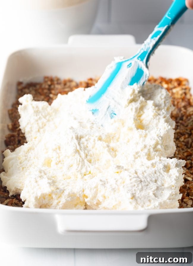 A blue spatula spreading a smooth layer of cream cheese and cool whip fluff over the baked pretzel crust in a baking dish.
