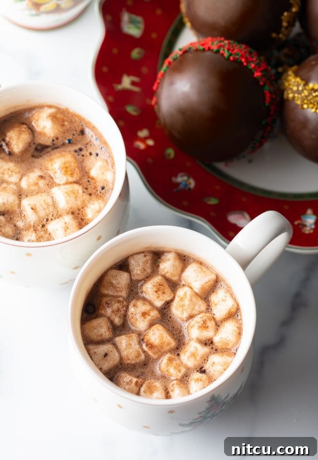 Two mugs of hot cocoa in Christmas mugs with plate of chocolate balls on festive plate in the background.