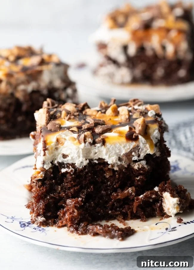 Close-up view of a chocolate poke cake generously topped with whipped cream, caramel and chocolate sauce, and scattered with chopped Heath bars, promising a rich and indulgent experience.
