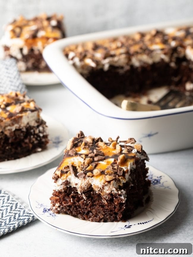 A slice of Better Than Sex chocolate poke cake, generously topped with whipped cream and caramel sauce, is beautifully presented on a white and blue plate, with the full baking dish and a spatula blurred in the background, creating an enticing scene.