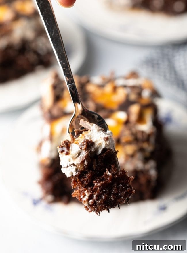 A fork lifts a generous bite of chocolate cake, showcasing the distinct layers of cake, rich caramel, fluffy whipped topping, and crunchy toffee candy bar pieces, promising a delightful texture and flavor combination.
