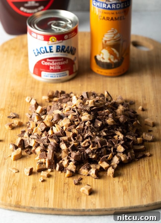 A wooden cutting board holds a pile of freshly chopped toffee bars. In the soft-focus background, a can of sweetened condensed milk and a bottle of caramel sauce are visible, highlighting the key ingredients for the cake's decadent topping and soak.