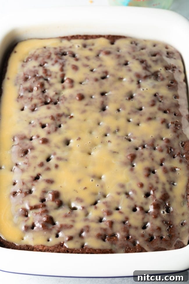 A stream of rich caramel sauce is poured evenly over the surface of the poked cake, ensuring that every hole is filled and the cake is thoroughly saturated for maximum flavor and moisture.
