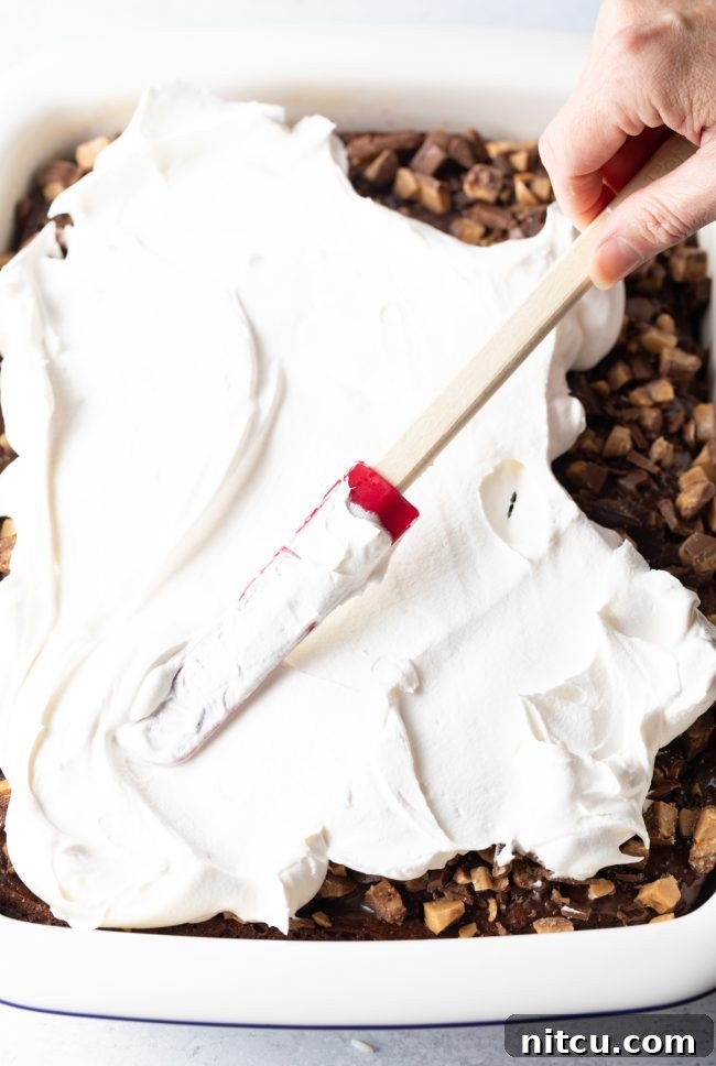 A hand is shown carefully adding a smooth, thick layer of whipped topping over the chopped candy bars on the cake, creating a creamy and inviting surface.