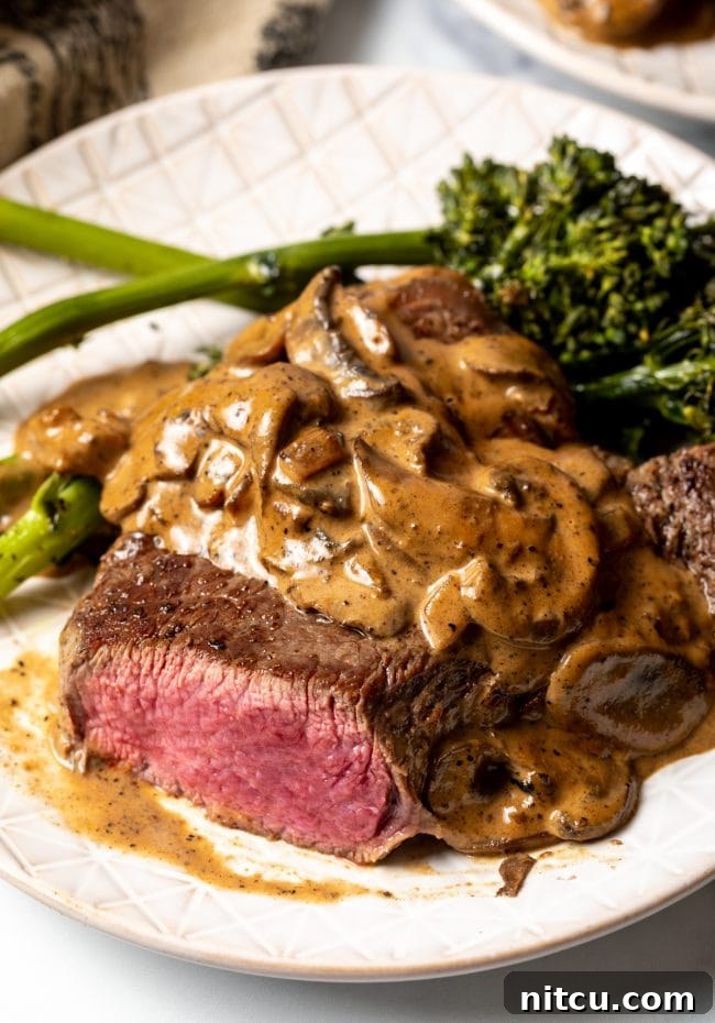 Close-up view of a perfectly sliced steak on a white plate, generously topped with a rich and creamy mushroom sauce, highlighting its tender interior.