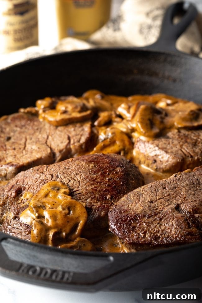Four perfectly cooked steaks nestled in a luscious mushroom cream gravy within a cast iron skillet, ready to be served.