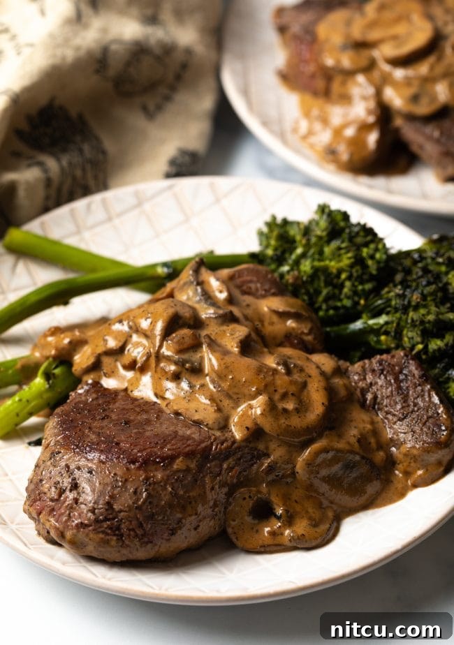 A thick, perfectly cooked Steak Diane on a white plate, lavishly covered with a luscious mushroom cognac gravy, with a side of vibrant broccolini.