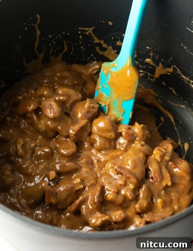 A blue spatula actively stirring pecan halves into freshly made, shimmering caramel inside a large pot.