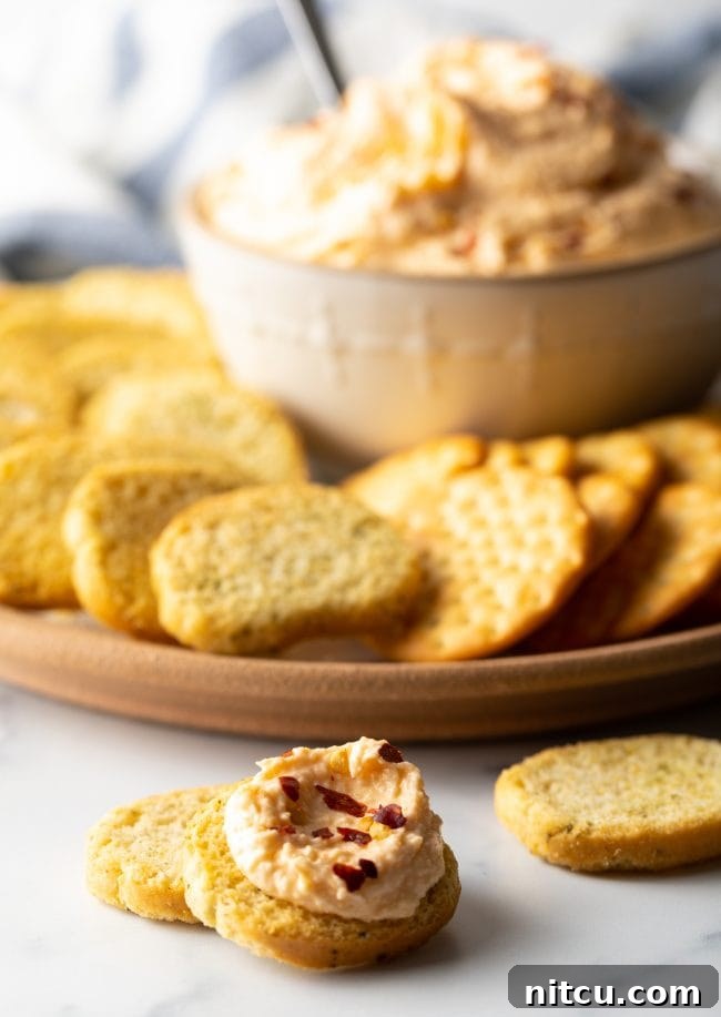 Pieces of sliced bread, one topped with smoked cheese dip and platter with crackers and bowl of spread in the background.