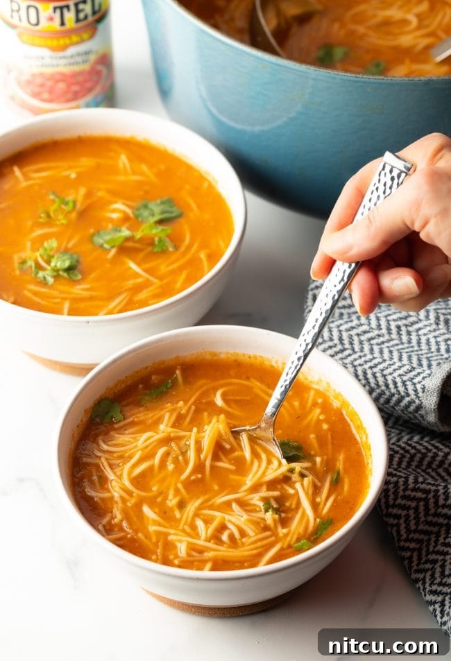 two bowls of sopa de fideo with a pot of soup in the background, foreground shows a hand holding a spoon scooping a bite of soup
