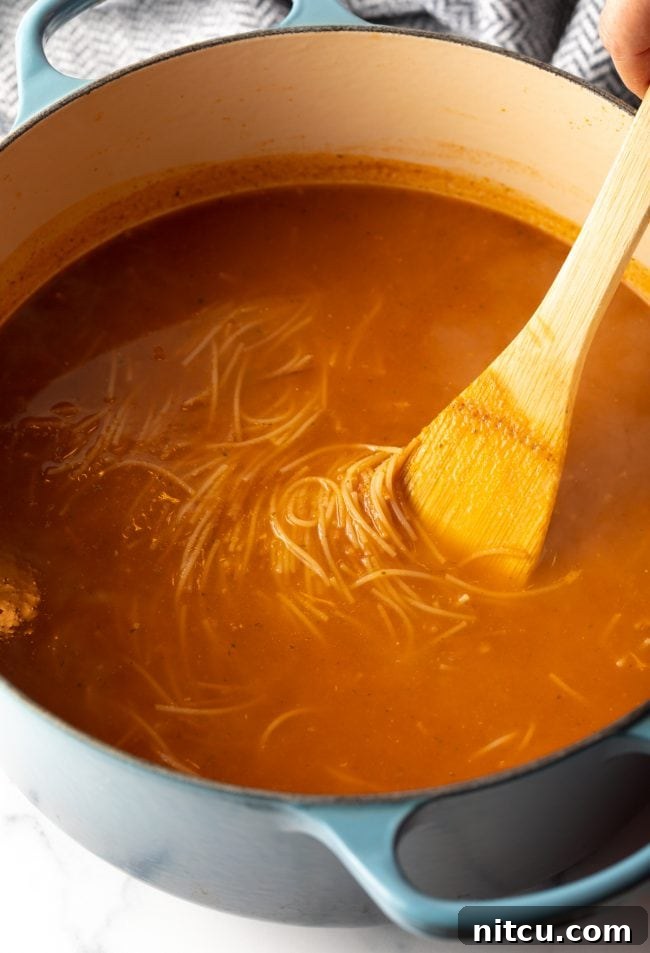 wooden spoon stirring Mexican tomato soup with noodles in a large pot, illustrating the cooking process