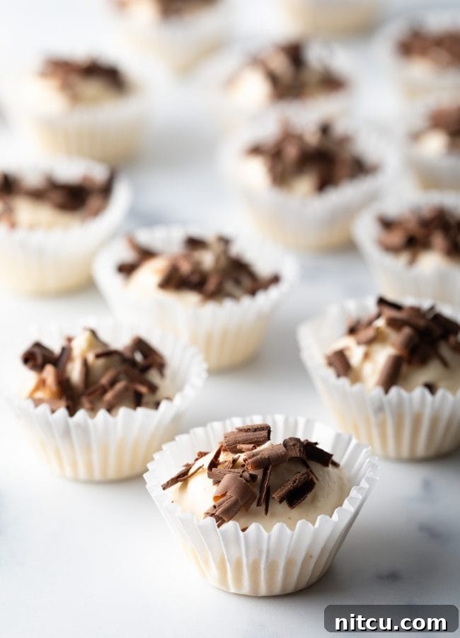 Close-up view of several no-bake keto cheesecake bites in small paper cups, adorned with delicate chocolate shavings, resting on an elegant marble background. A perfect low-carb dessert.
