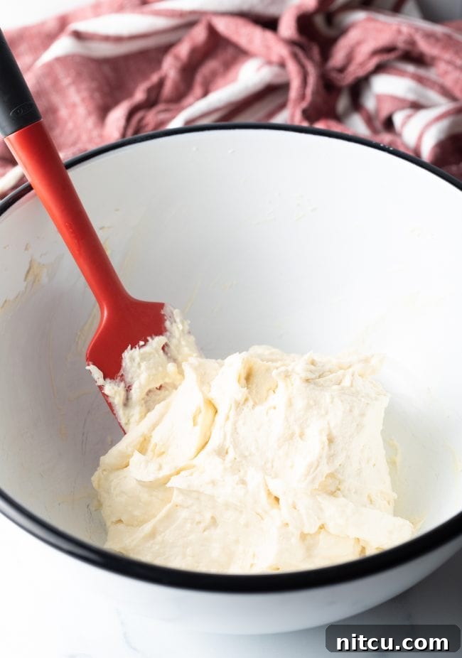 A large mixing bowl containing the creamy white cheesecake mixture, with a red silicone spatula poised to combine the ingredients.