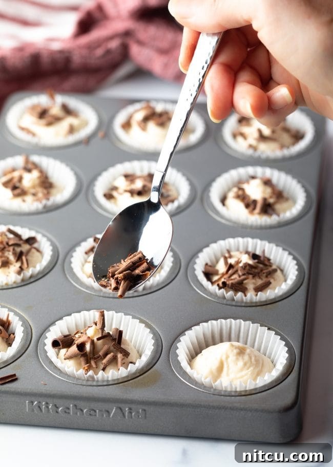 A hand using a spoon to delicately add thin shavings of dark chocolate as a topping to the no-bake cream cheese cheesecake bites, enhancing their visual appeal and flavor.