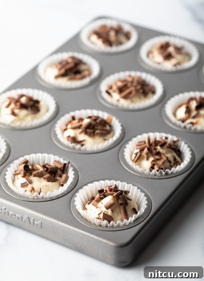 A mini muffin tin neatly arranged with white paper liners, each filled with a perfectly portioned keto cheesecake bite, generously topped with chocolate shavings, ready for chilling or serving.